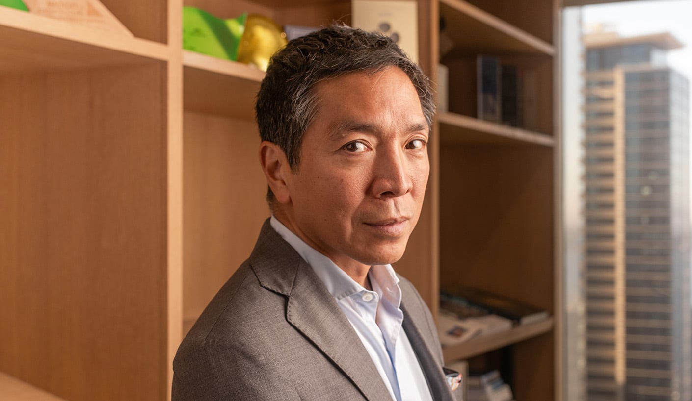 Portrait of a man in a suit, facing the camera with shelves in the background displaying decorative items and books, representing a professional environment.
