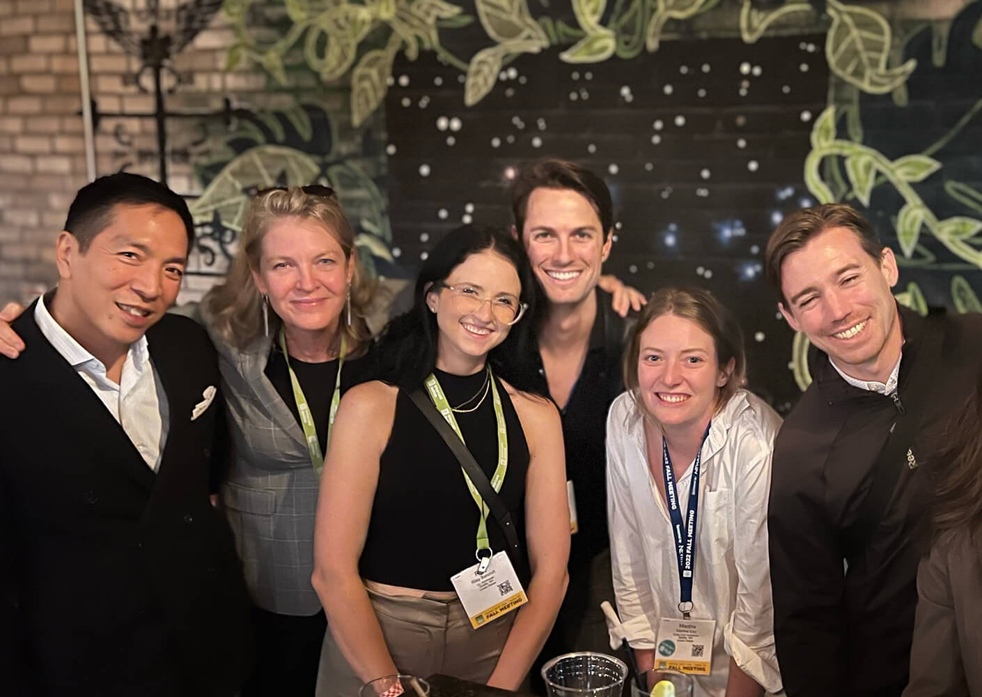 Group of six people smiling together at an event, with name tags visible, in front of a decorated wall with greenery.