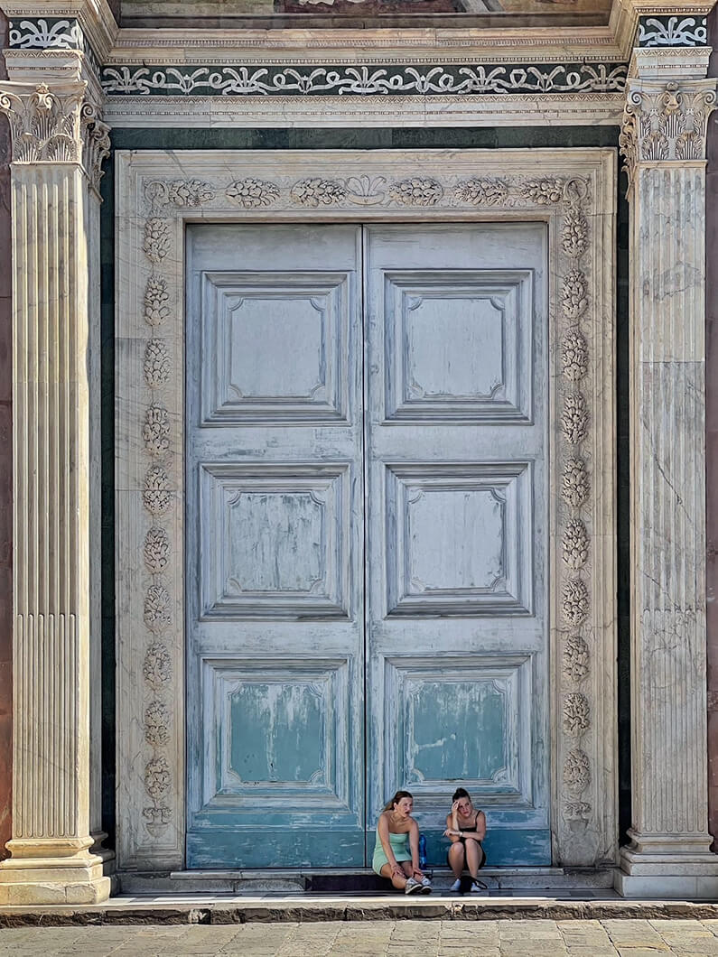 Two women sitting on steps in front of a large, ornate door framed by decorative columns and detailing. Bright daylight illuminates the scene.