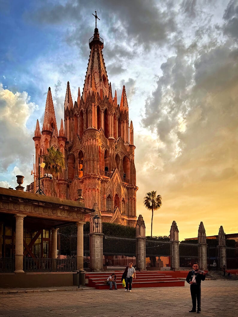 Church with intricate spires under a dramatic sky; people relax nearby, while a musician plays in a vibrant plaza.