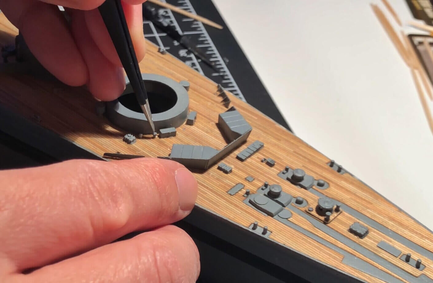 Close-up of a hand assembling a detailed ship model, using a fine tool on textured wooden surface with various small components.