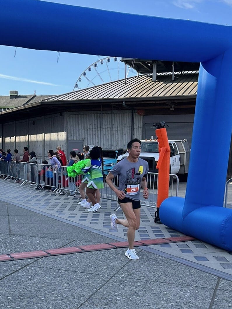 A runner crosses the finish line under a blue inflatable arch, with spectators and cheerleaders in the background near an event venue.