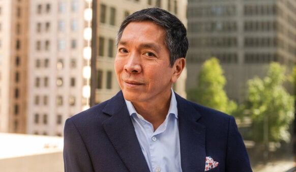 A man in a dark suit and light blue shirt smiles confidently, with buildings and greenery in the background.